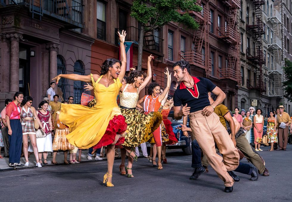 Two lines of dancers, one of women in colorful dresses and the other of men in slacks, lean toward each other on an NYC street